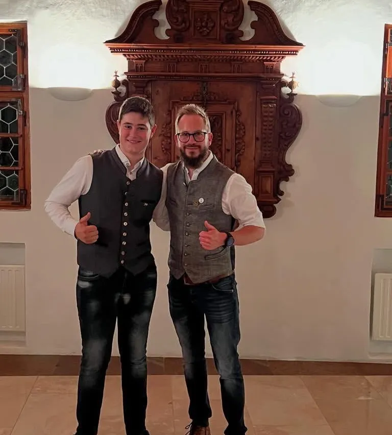 Zwei Männer stehen drinnen nebeneinander und geben Daumen hoch. Einer trägt eine Weste und der andere einen Bart. Hinter ihnen steht ein Holzschrank und zwei Lampen.