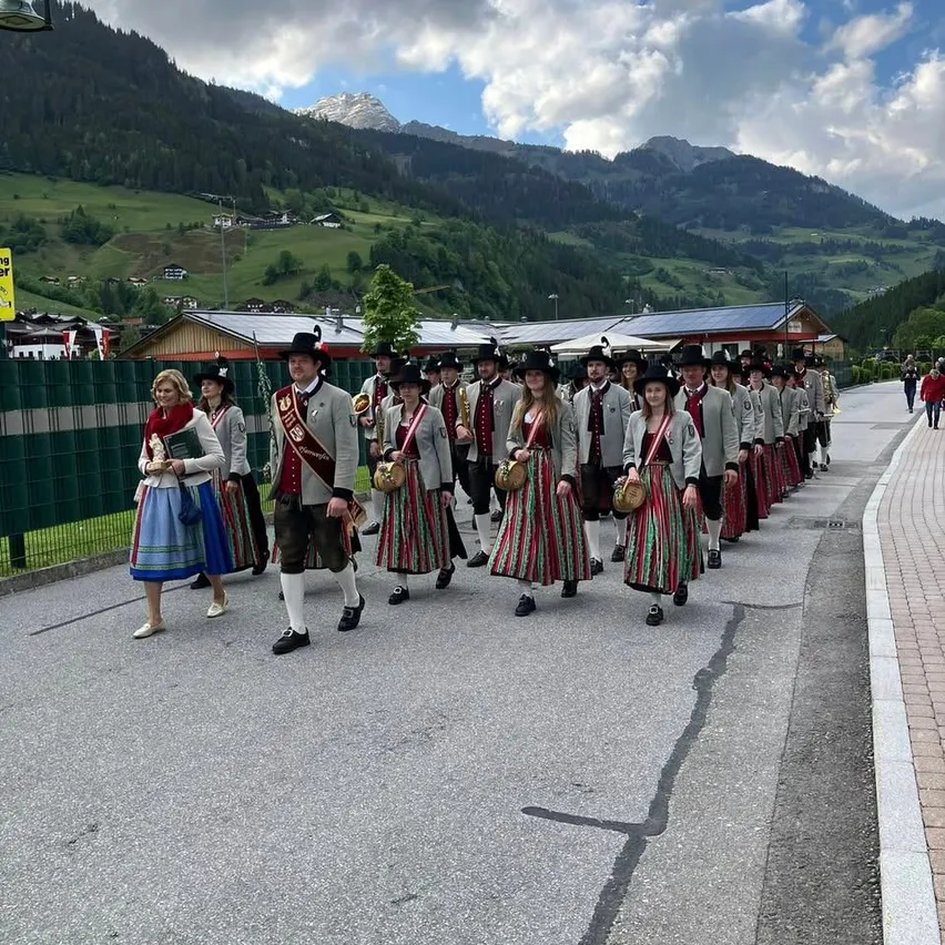 Eine Blaskapelle in traditioneller österreichischer Tracht geht auf einer Straße in den Bergen. Die Gruppe wird von einer Frau angeführt, und die Berge sind im Hintergrund.