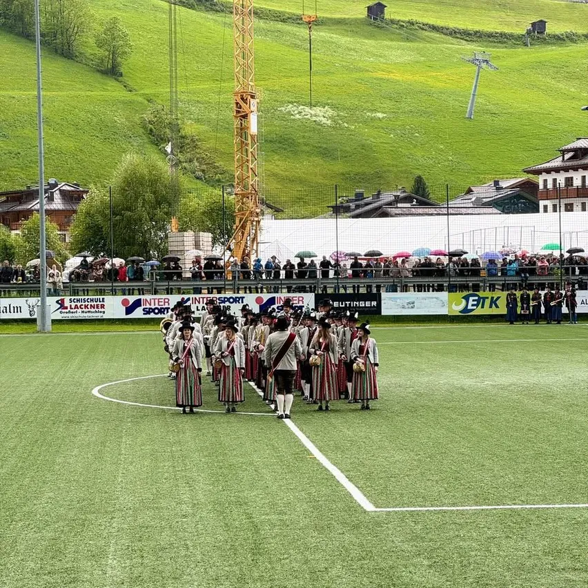 Eine Marschkapelle in traditioneller Kleidung tritt auf einem grünen Feld auf, während eine Menschenmenge mit Regenschirmen im Hintergrund zuschaut. Der Hintergrund zeigt einen Zaun mit Werbung, einen Turm und Häuser auf einem Hügel.