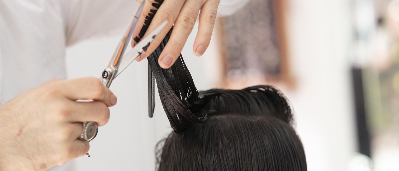 Eine Person schneidet mit einer Schere die Haare einer Frau. Die Frau hat lange, dunkle Haare. Die Schere ist silber und orange. Die Frau trägt ein weißes Oberteil.