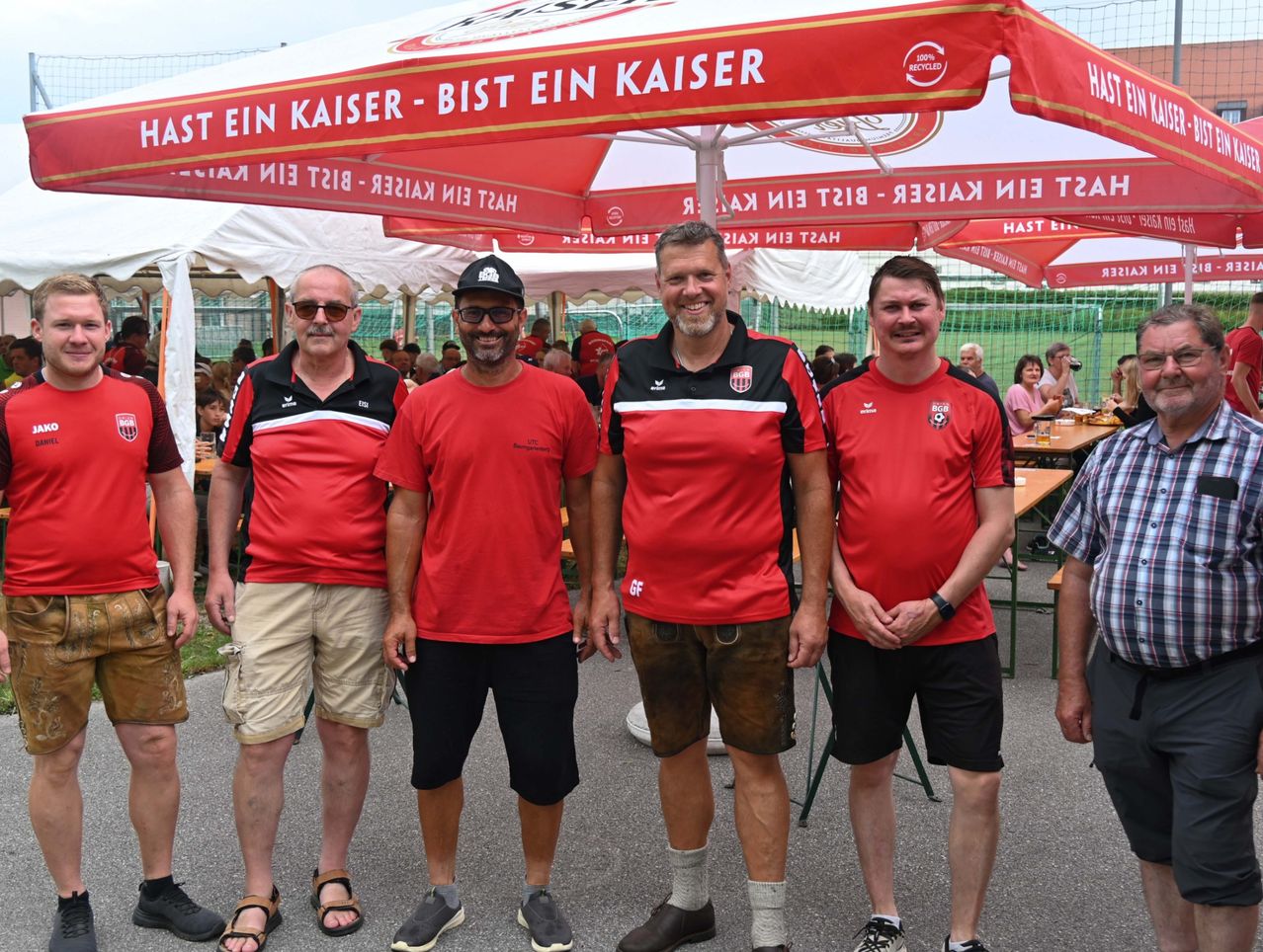 Vier Männer in roten Hemden und Shorts stehen unter einem roten und weißen Zelt mit einem Slogan darauf. Sie lächeln für die Kamera. Hinter ihnen stehen Tische mit Essen und sitzende Menschen.