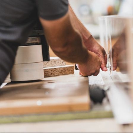 Eine Person benutzt ein Werkzeug, um ein Stück Holz auf einem Tisch zu schneiden. Sie trägt ein graues Shirt.