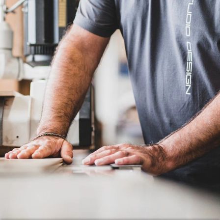 Ein Mann in einem grauen T-Shirt arbeitet mit seinen Händen an einem Tisch. Der Tisch hat eine glatte Oberfläche. Hinter ihm steht eine Maschine, möglicherweise eine Drehbank.