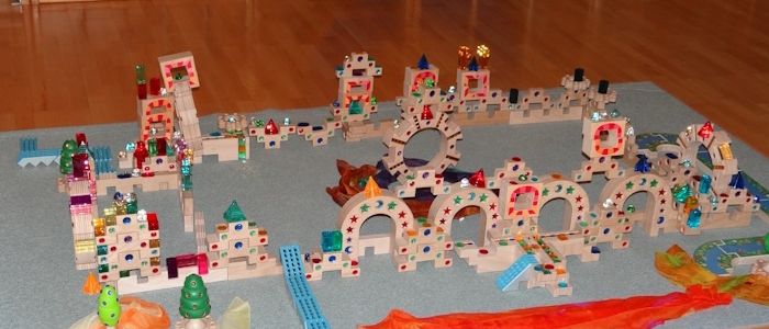 Ein Holzboden zeigt eine farbenfrohe, komplizierte Anordnung von Holzklötzen, die einer Miniaturstadt ähnelt. Verschiedene Gebäude, Bäume und ein gewundener Weg sind aus farbigen Blöcken gefertigt. Ein orangefarbener Stoff ist auf dem Boden ausgebreitet.