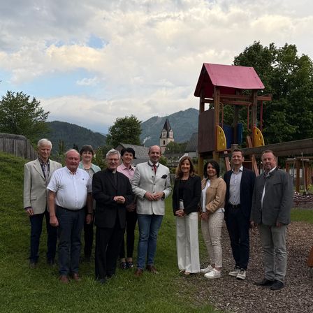 Eine Gruppe von Menschen steht vor einem Spielplatz. Sie sind formell gekleidet und scheinen für ein Foto zu posieren.