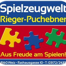 Ein blau-roter Schild mit weisser Schrift zeigt 'Spielzeugwelt Rieger-Puchebner', ein Puzzlestück-Logo und Kontaktdaten unten.