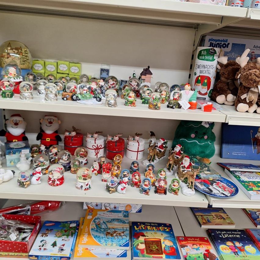 Ein weißer Regal mit mehreren Reihen von Weihnachtsschmuck und Spielzeug, darunter Santa-Figuren, Schneekugeln und Plüschtiere. Es gibt auch Bücher, Puzzles und Geschenkboxen und einen grünen Weihnachtsbaum auf der rechten Seite.