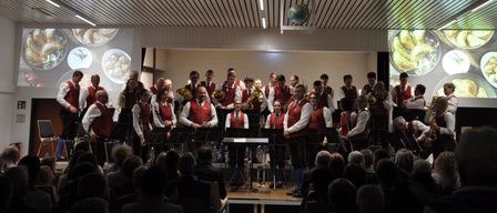 Eine Gruppe von Musikern, gekleidet in rote Westen und weiße Hemden, spielt auf der Bühne vor einem Publikum. Eine Erdkugel hängt von der Decke, und an den Wänden sind Bildschirme mit Essensbildern angebracht.