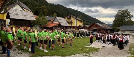 Eine Band in grünen und gelben Uniformen steht auf einem Schotterweg vor Häusern mit Bergen im Hintergrund.
