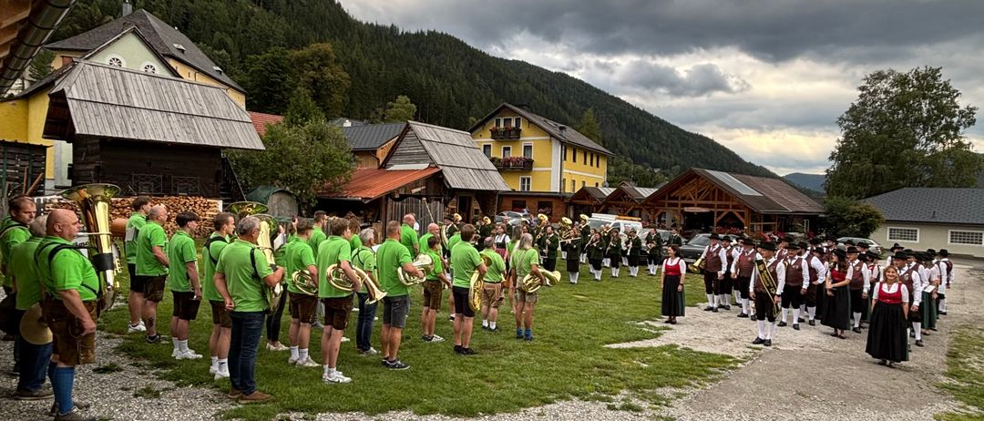 Eine Band in grünen und gelben Uniformen steht auf einem Schotterweg vor Häusern mit Bergen im Hintergrund.