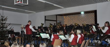 Ein Orchester spielt auf einer Bühne mit verschiedenen Instrumenten, darunter Blech- und Holzblasinstrumente. Das Publikum sitzt in Stühlen und beobachtet aufmerksam die Aufführung. Ein Dirigent leitet die Musiker.