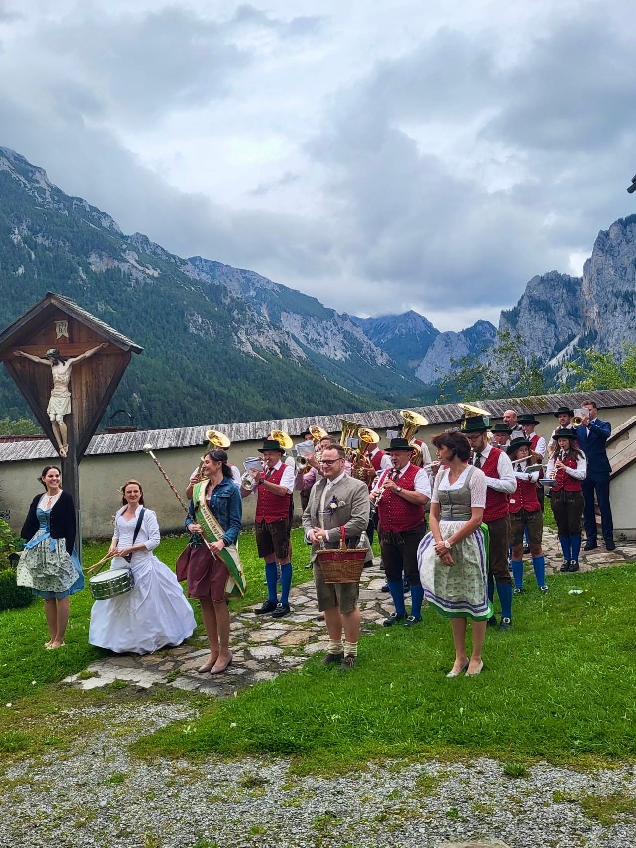 Eine Gruppe von Musikern steht in traditioneller Kleidung, mit Bergen im Hintergrund. Einige spielen Blechblasinstrumente, andere haben Flöten und eine Frau hält eine Trommel.