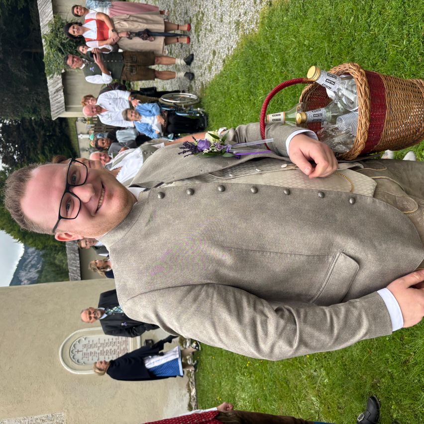 Ein Mann in einem Anzug, der einen Korb mit Flaschen hält, liegt auf einer Rasenfläche. Hinter ihm stehen Menschen in traditioneller Kleidung, einer davon im Rollstuhl. Im Hintergrund befindet sich ein Gebäude.