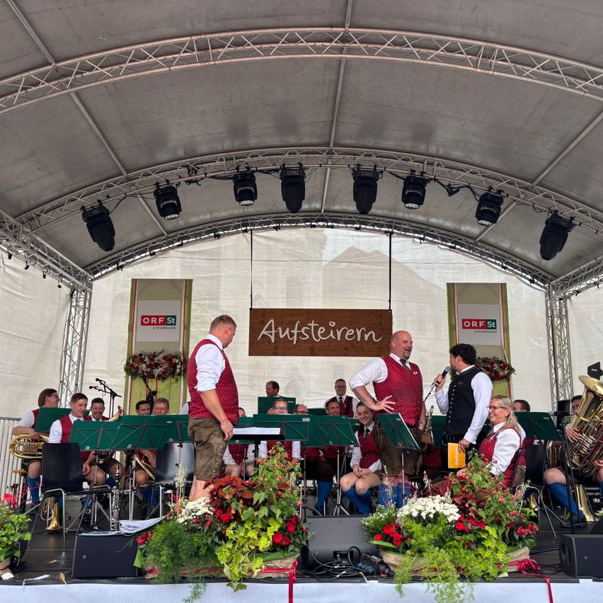 Ein Blasorchester spielt auf der Bühne mit großen Blumenarrangements, unter einem Metallrahmen mit Scheinwerfern und einem Banner mit der Aufschrift Aufsteirem.