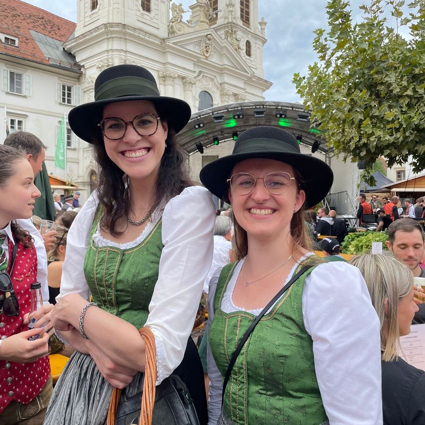 Zwei Frauen in traditionellen grünen Kleidern und Hüten lächeln in die Kamera und stehen vor einer großen Menschenmenge, mit einer Kirche und einem grünen Baum im Hintergrund.