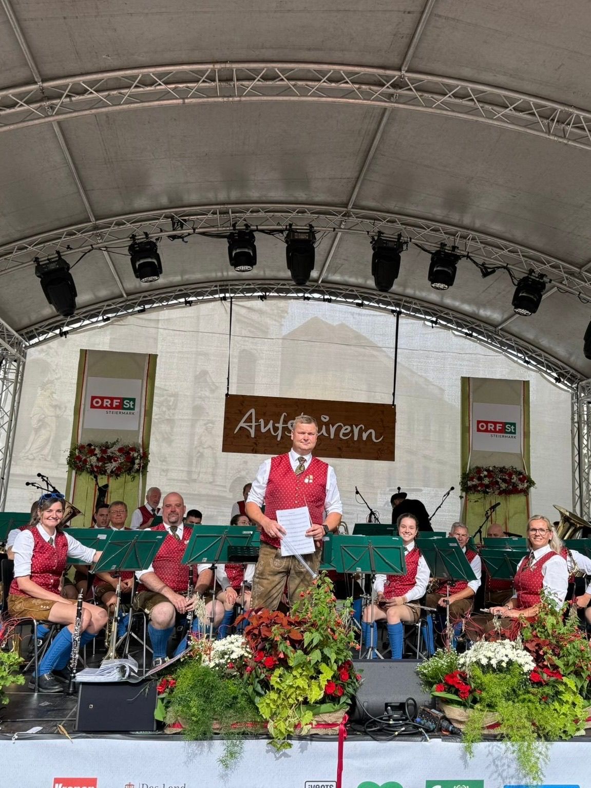 Ein Orchester auf der Bühne unter einem Zelt mit Musikern in traditioneller deutscher Kleidung. Ein Mann steht vorne und hält ein Papier, während andere Instrumente spielen. Blumendekorationen schmücken die Bühne.