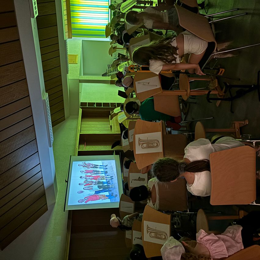 Eine Vogelperspektive eines Klassenzimmers mit Schülern an Tischen sitzend, einige schauen auf einen Bildschirm mit einer Gruppenfoto. Der Raum hat Holzwände und eine Projektionswand.