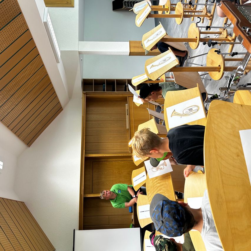 Mehrere Kinder sitzen in einem Klassenzimmer um Musikinstrumente herum. Ein Lehrer in Grün steht hinter ihnen, und die Kinder haben Blätter mit Musiknoten auf ihren Schreibtischen.