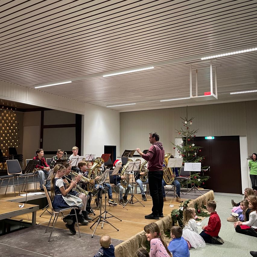 Eine Kinderband spielt Blechblasinstrumente auf der Bühne in einer Halle mit einem Weihnachtsbaum. Ein Mann steht davor und mehrere Kinder sitzen auf dem Boden und zuschauen.