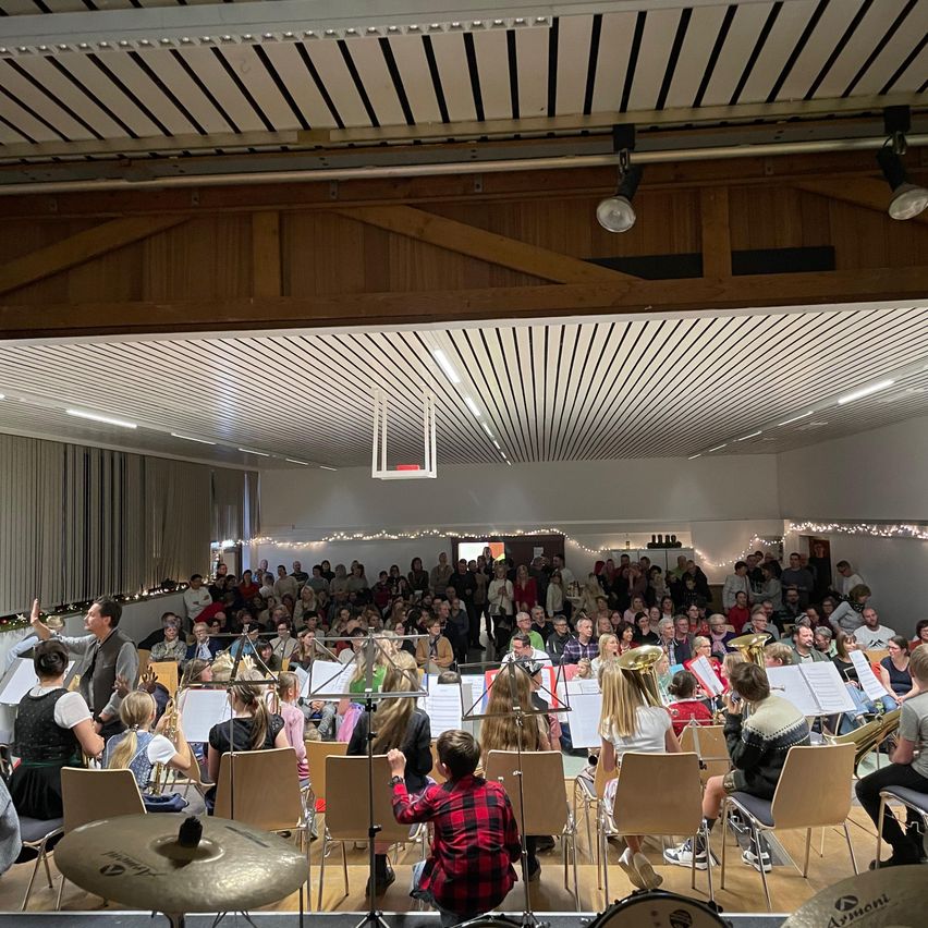 Ein großes Publikum füllt einen Raum mit Holzbalken, während eine Brass-Band auf einer Bühne mit Musikinstrumenten und Notenblättern auftritt.