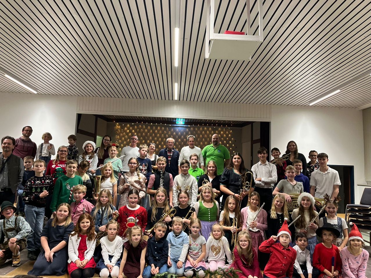 Eine Gruppe von Kindern und Erwachsenen posiert für ein Foto in einem Raum. Einige von ihnen halten Musikinstrumente wie Trompeten, Posaunen und Flöten. Es sind auch Erwachsene im Raum. Der Raum hat weiße Wände und eine Decke mit schwarzen und weißen Streifen. Es hängen auch Lampen an der Decke.