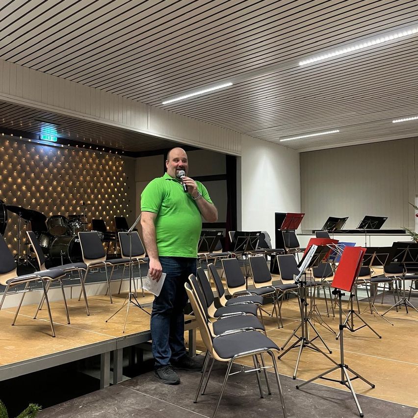 Ein Mann steht auf einer Bühne in einem Musiksaal, hält ein Mikrofon in der Hand, um ihn herum sind Stühle und Instrumente aufgestellt.