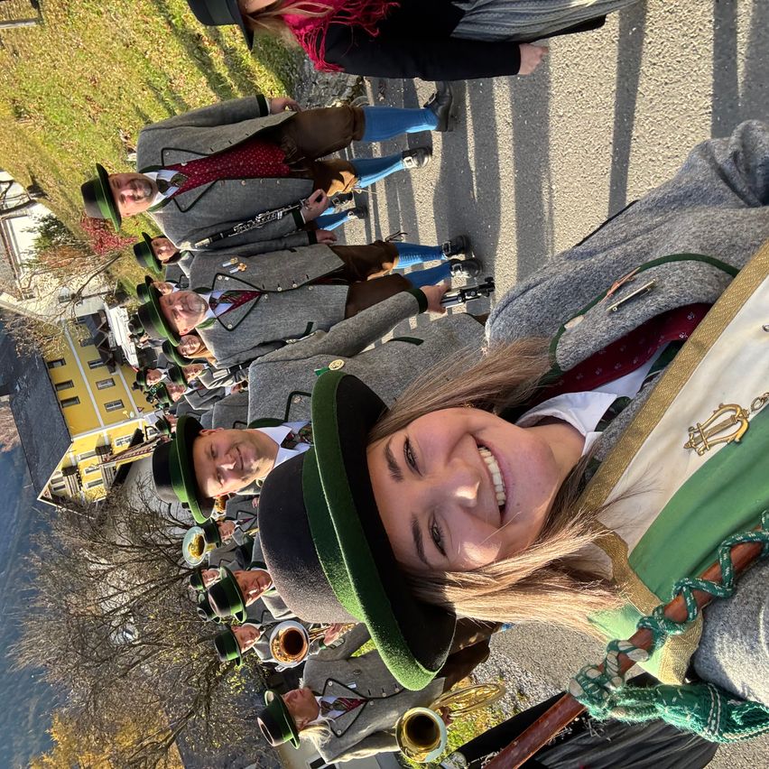A group of people dressed in traditional German attire, with green hats, smile for a photo. They are standing on a road with a yellow building in the background.