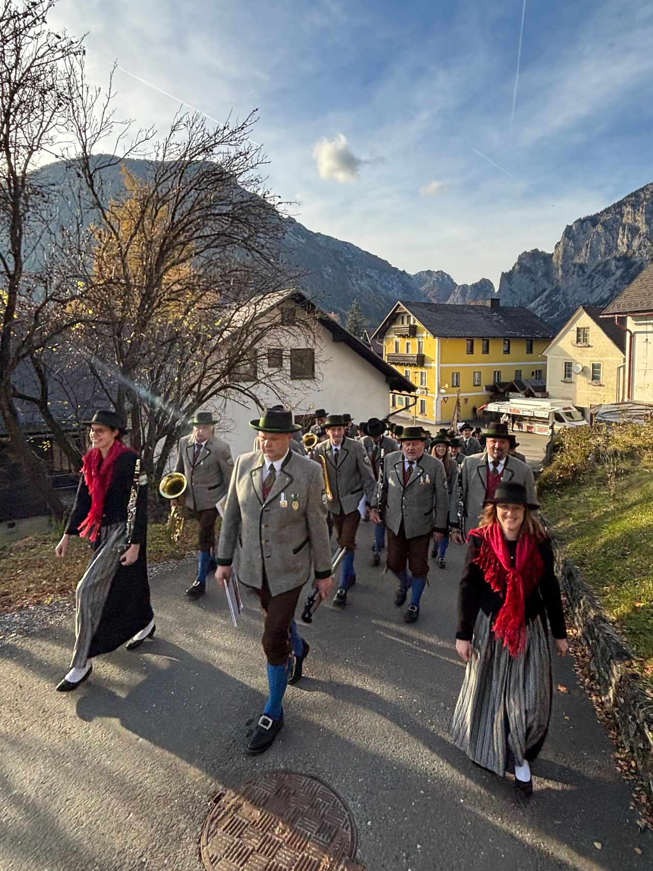 A marching band, dressed in traditional attire, is walking down a village road, accompanied by a female member, with mountains in the background.