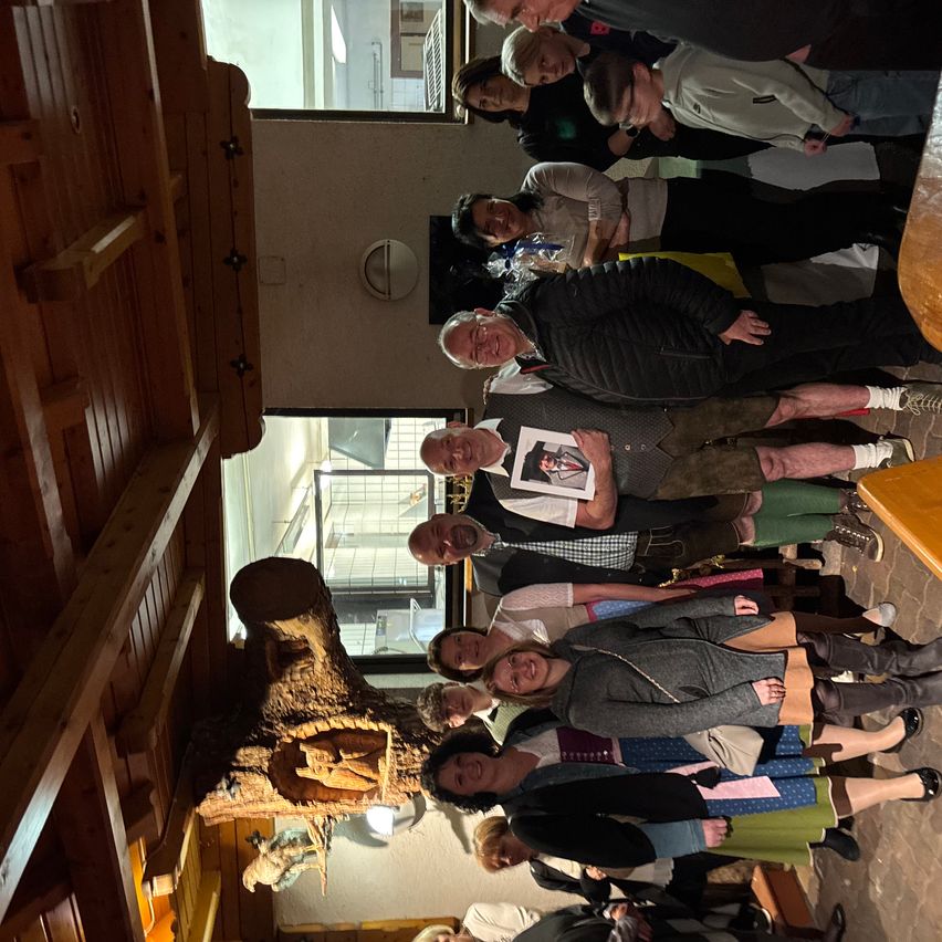 A group of people stand together in a room, some smiling and looking at a carved wooden log. The room has wooden walls and windows with a view of the outside.
