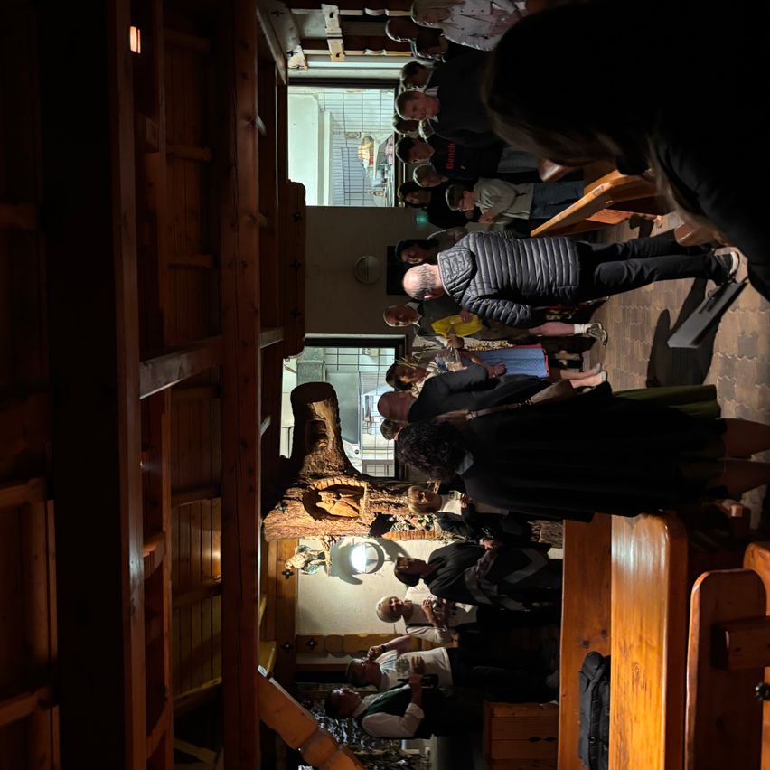 A group of people are gathered in a room with wooden walls, benches, and a carved tree trunk. Some are standing, while others are sitting and chatting. A window offers a view of a building.