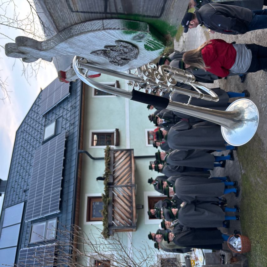 A group of people in traditional clothing gather around a silver trombone near a house with solar panels and a wooden balcony.