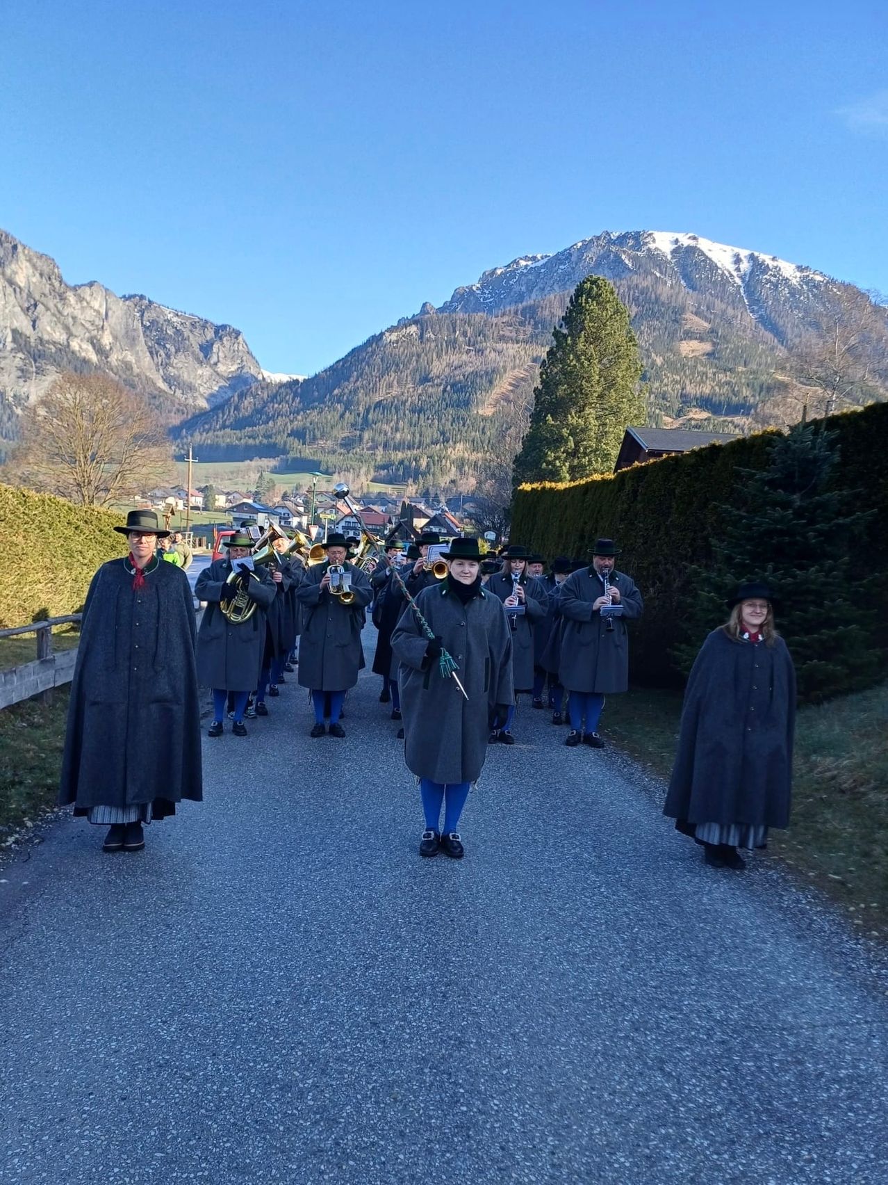 A marching band in traditional attire is walking in a line on a road with mountains in the background.