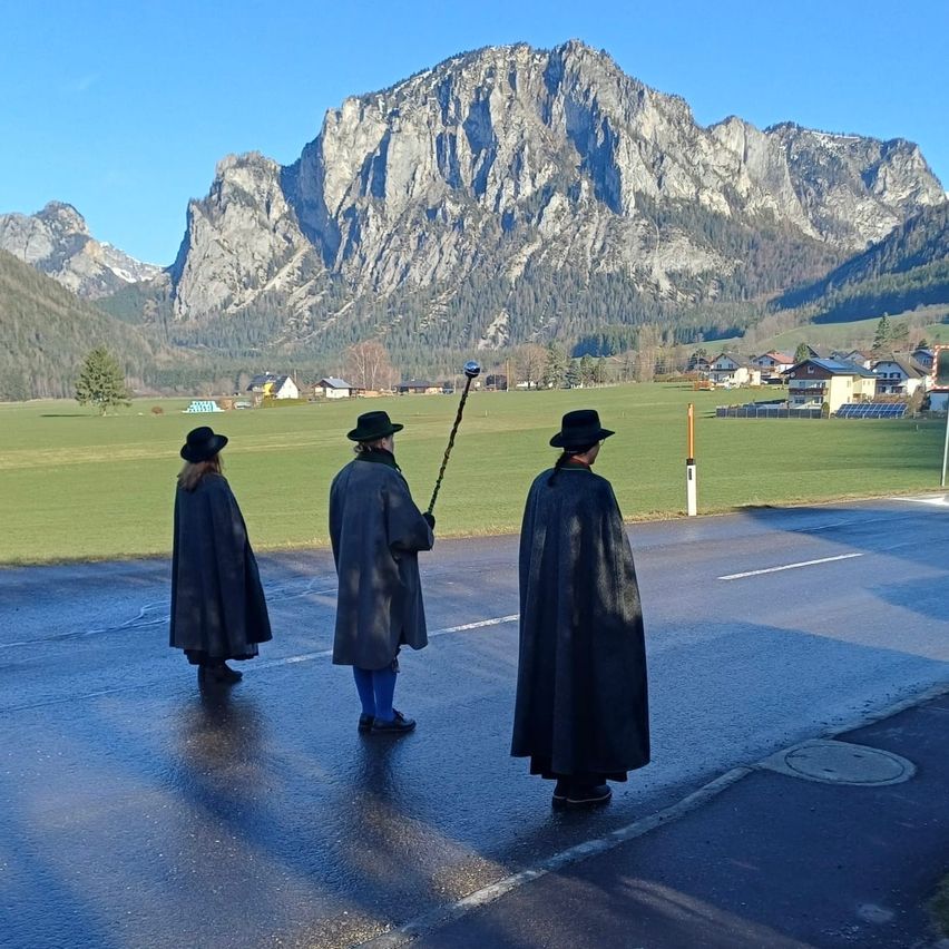 Three individuals in matching cloaks and hats stand in a wet road with mountains in the background. They are holding a pole with a flag.