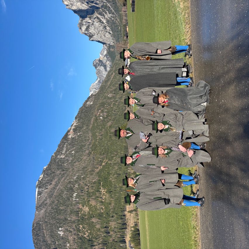 A group of people dressed in gray coats and hats pose for a photo on a mountain road. The background shows a mountainous landscape.