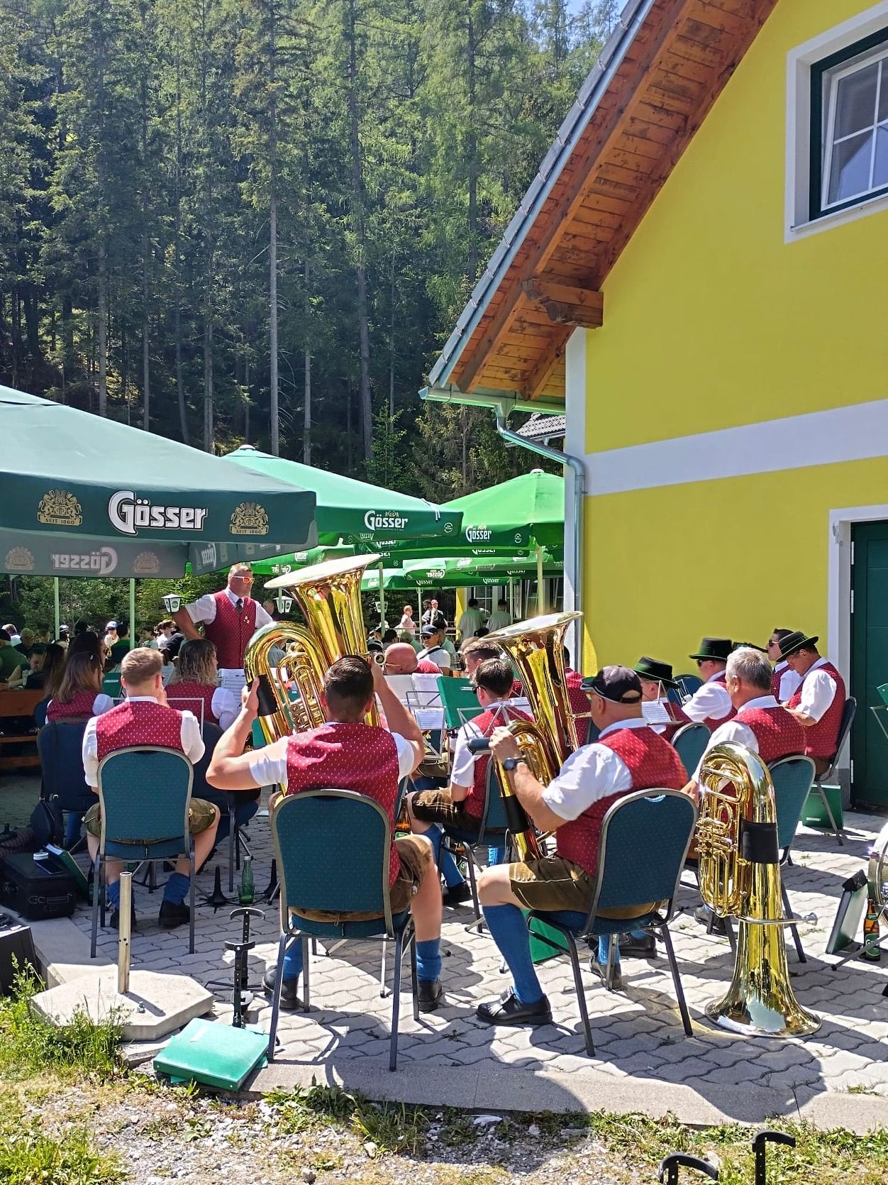 A group of musicians plays brass instruments outside. They are seated in front of a yellow building, with green umbrellas in the background.