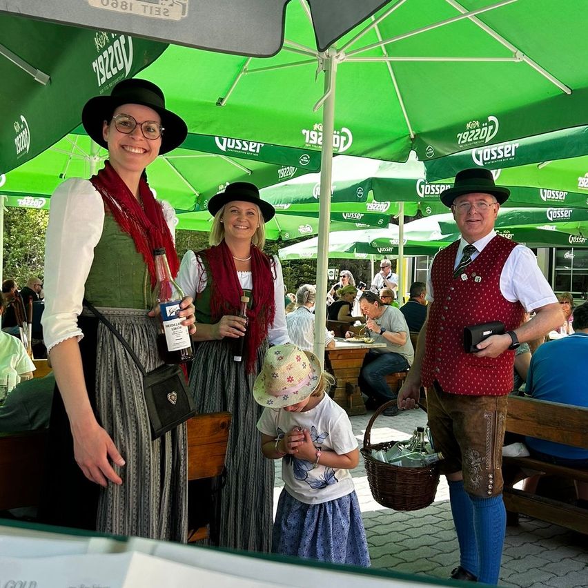 Three people are standing under a green umbrella. The two women are wearing traditional Bavarian clothing. The man is wearing a red vest and holding a basket. A child is standing in front of them. Behind them, several people are sitting on benches, some of whom are eating.