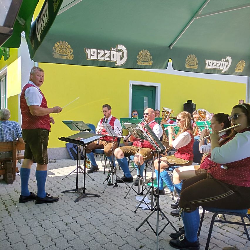 A band performs under a green umbrella, with a conductor in front. Members sit and play instruments like trumpets and tubas. They wear traditional German attire.