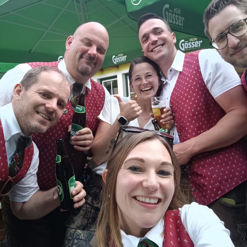 Five people wearing red vests are posing for a picture. They hold bottles and a glass of beer. The woman in the middle smiles at the camera. They are under a green umbrella.