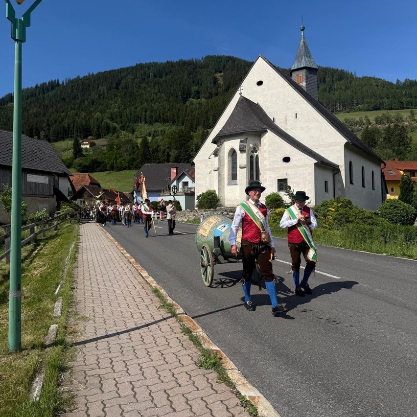 A procession in traditional attire walks down a street with a church in the background. Two men lead the procession.