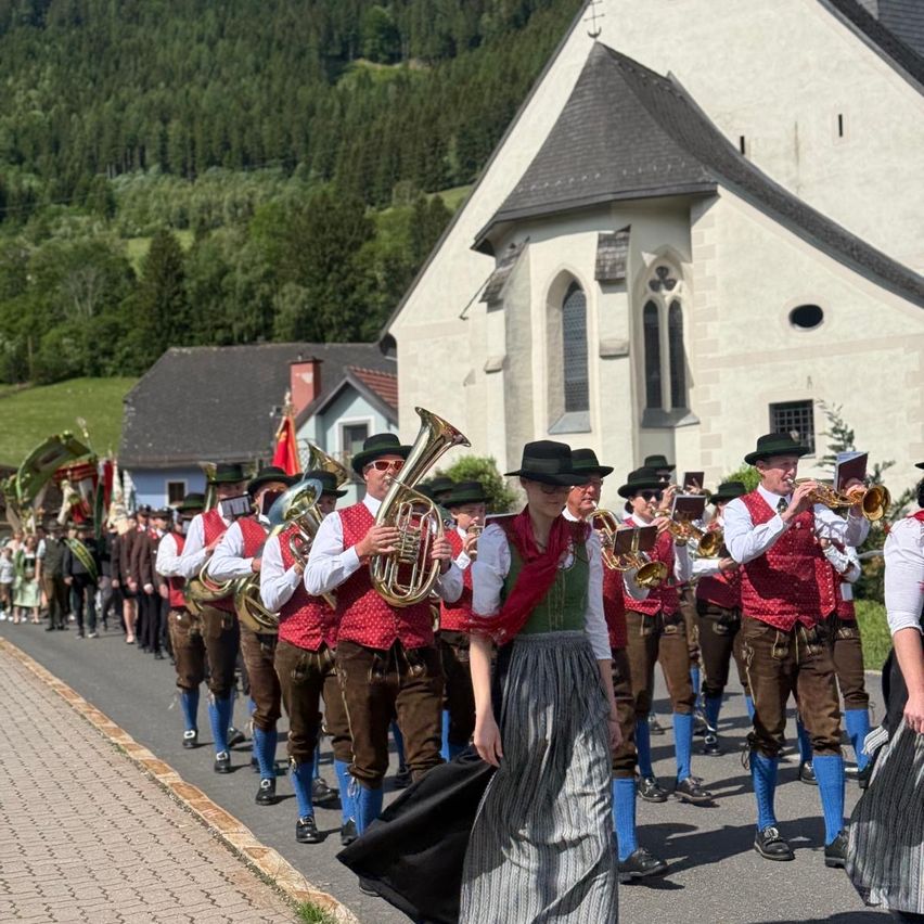 A marching band with brass instruments walks on a road in front of a church, with spectators on the side. The band members wear traditional costumes, and the sky is clear.