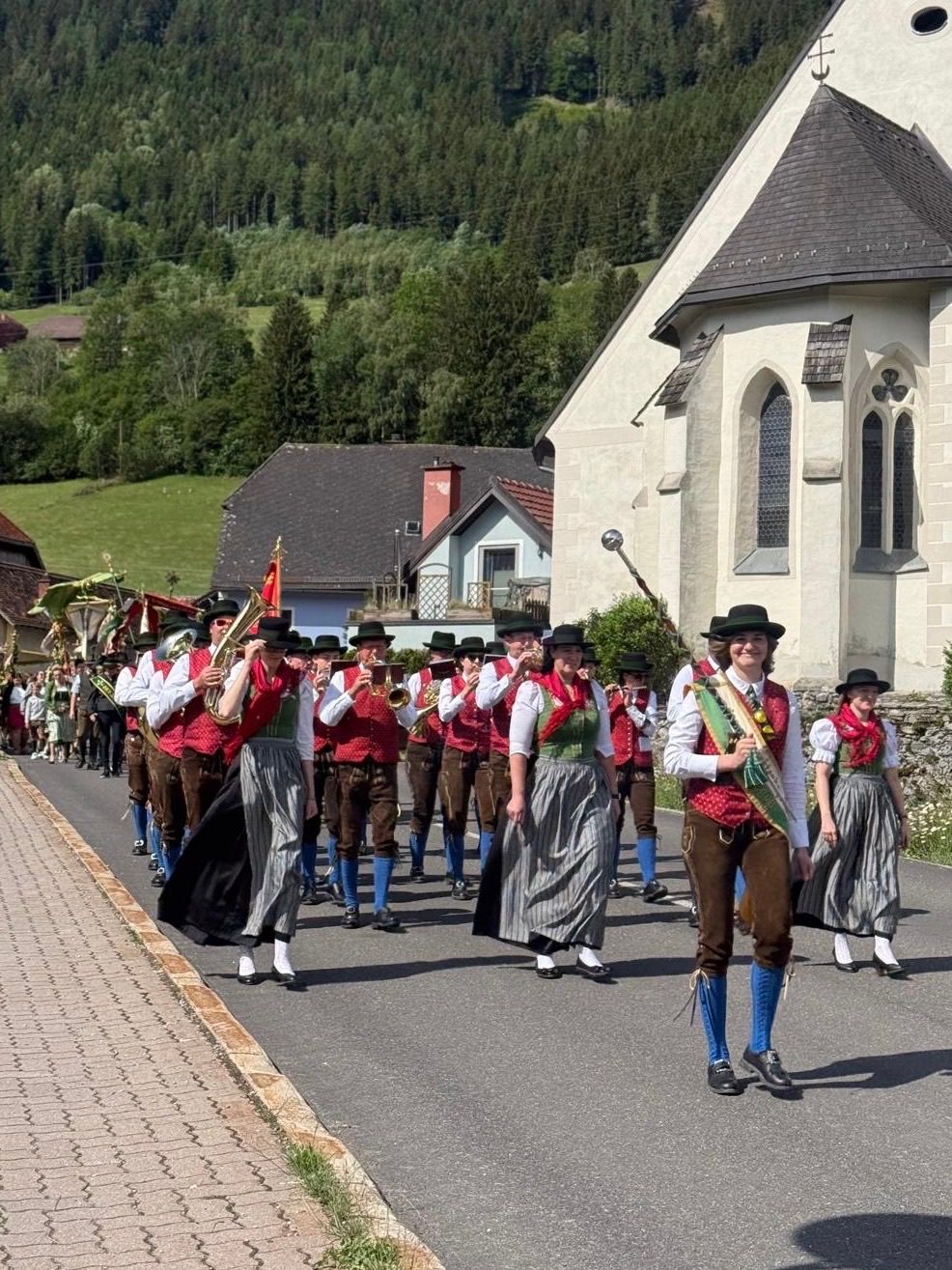 A brass band of men and women in traditional Bavarian clothing marches down a street in a village.