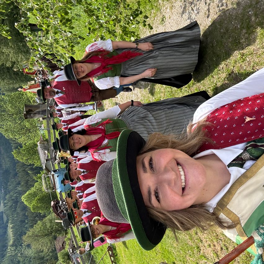 A group of people dressed in traditional Bavarian attire, standing in a field. They appear to be posing for a photo, with one woman smiling brightly in the foreground.