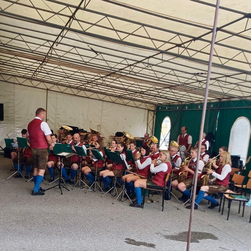 A brass band in traditional attire is performing under a white tent. Several musicians play trumpets, trombones, and saxophones, while others sit and play their instruments.
