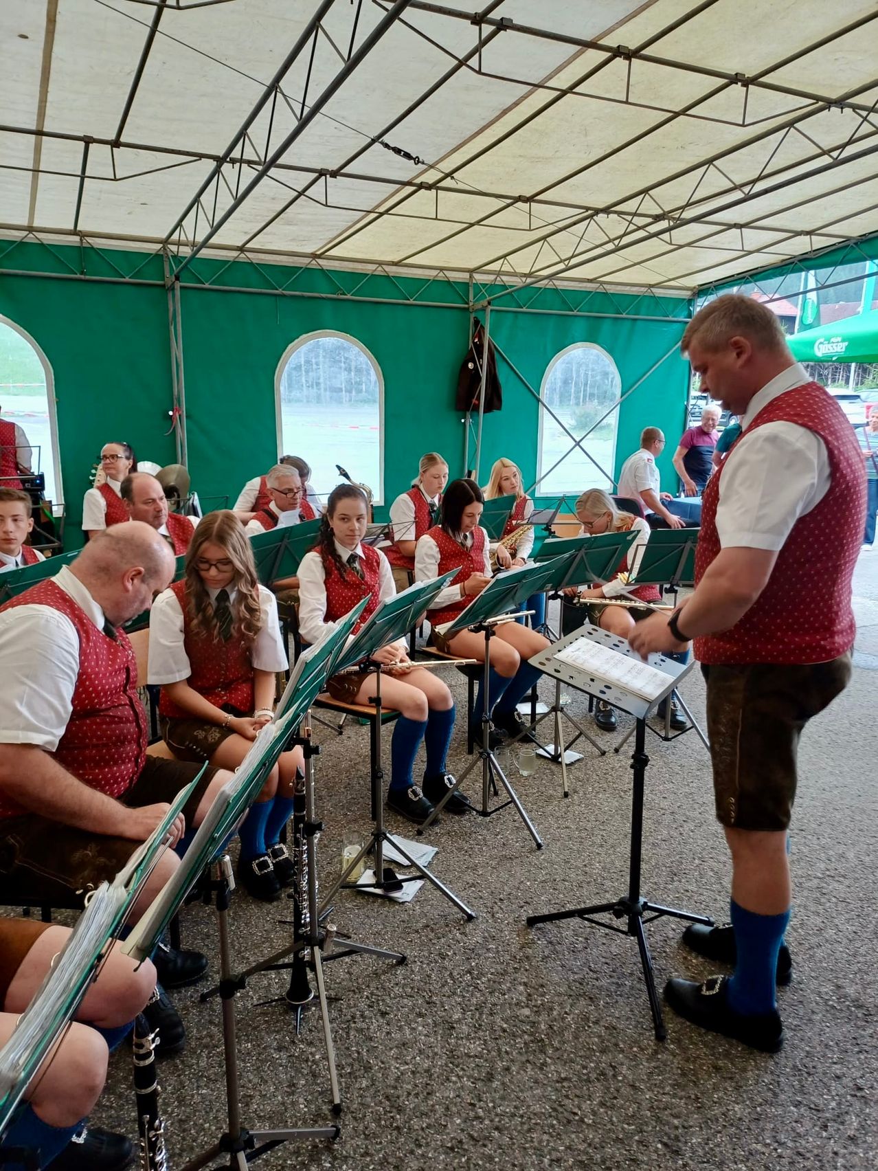 A band under a tent practices with musical instruments; musicians sit while a man stands by them.