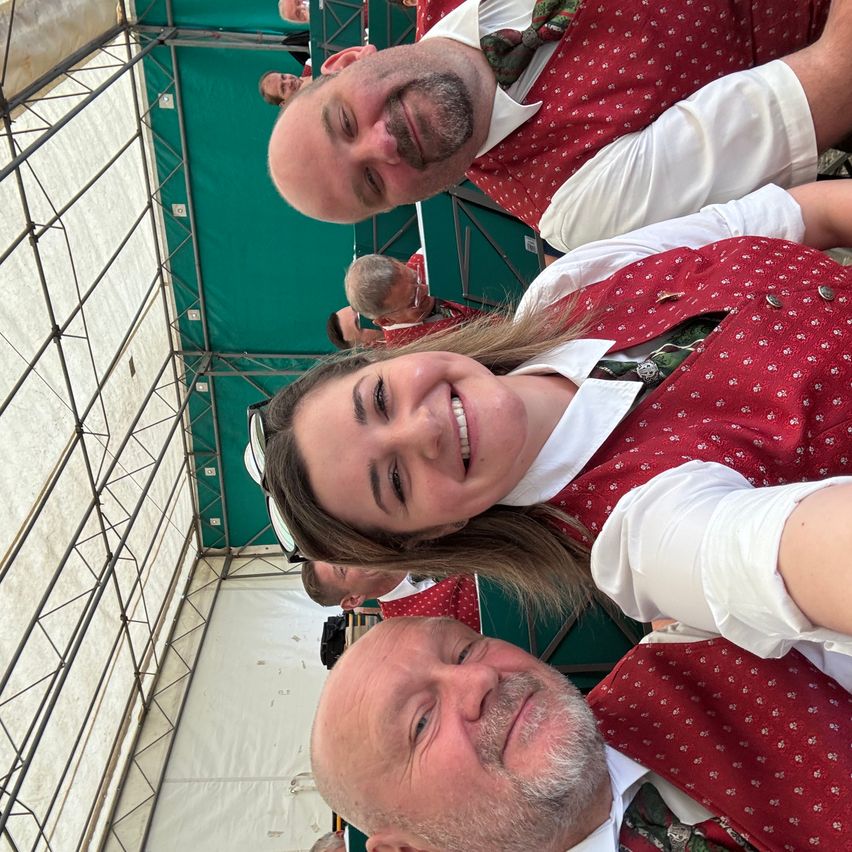 Several people in festive attire are smiling at the camera. A woman with a red vest is in the center. Two men are on either side, also wearing red vests. Behind them, there is a green tent.