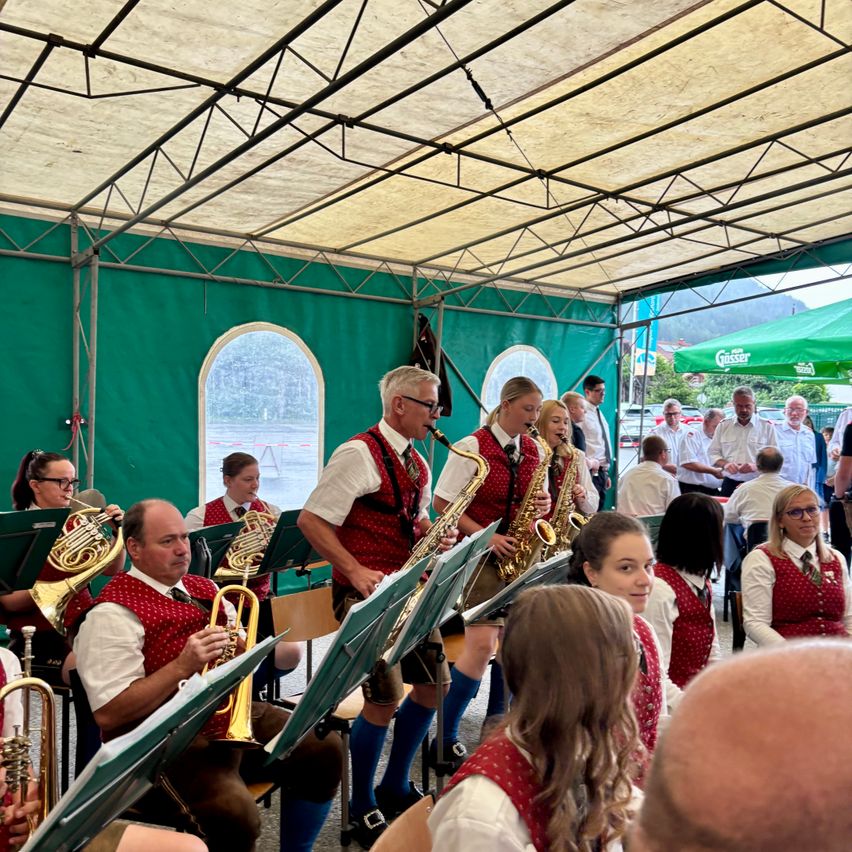 A brass band plays under a tent, with musicians wearing red vests and some in traditional clothing. Spectators watch from chairs, and others stand behind them.