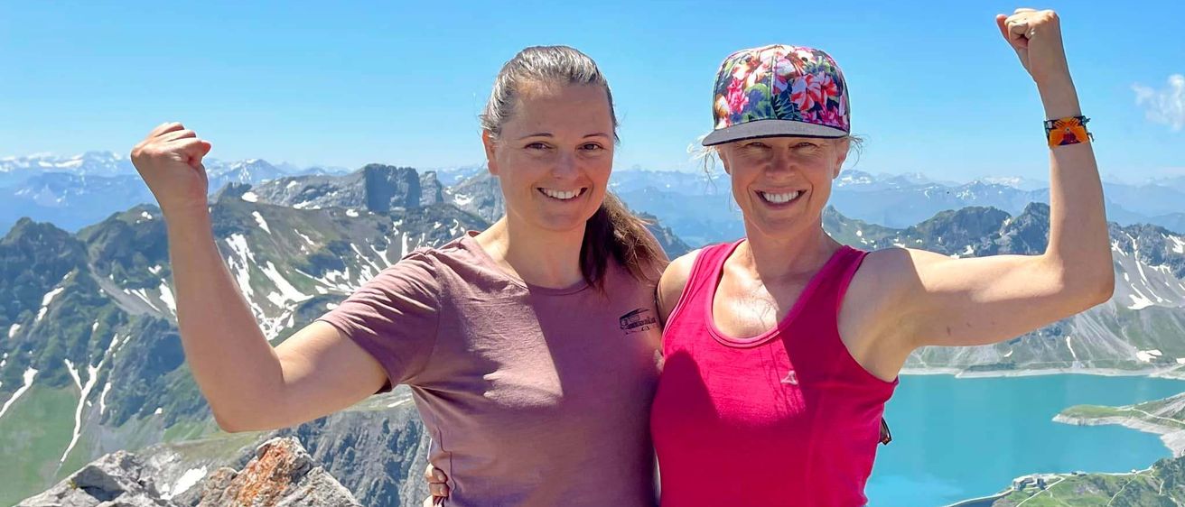 Zwei Frauen stehen auf einem Berggipfel, Arme erhoben, lächeln. Hinter ihnen ein ruhiger See und schneebedeckte Berge unter einem klaren Himmel.