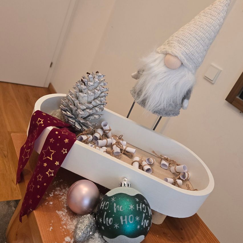 Eine weiße Badewanne, gefüllt mit Weihnachtskugeln, darunter eine Tannenzapfen, ein Gnom und ein rotes Band mit Sternen, steht auf einem Holztisch mit Marmorplatte.