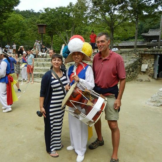 Ein Mann in einem bunten Hut und traditioneller Kleidung hält eine Trommel und steht neben einer Frau und einem Mann. Dahinter versammeln sich Menschen in traditionellen Kostümen auf Steinstufen. Bäume und ein Gebäude sind im Hintergrund.