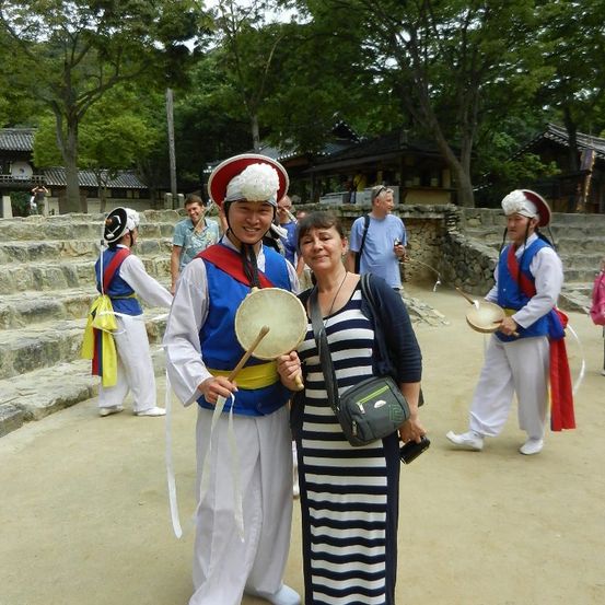 Ein Mann und eine Frau stehen lächelnd zusammen, in traditioneller koreanischer Kleidung, mit Instrumenten und einem Steingebäude im Hintergrund.